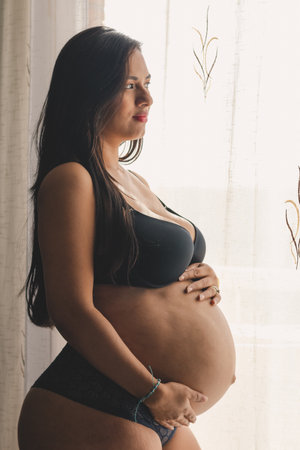 Pregnant young woman touching her belly in black underwear, looking at the horizon next to a window.の写真素材