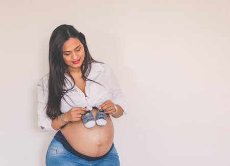Pregnant young woman holding small shoes on her belly, smiling and with copy space on a white wall.