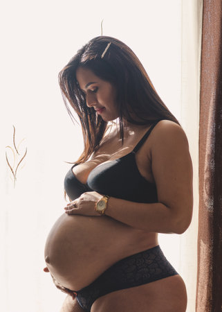 Young pregnant latin woman looking and touching her belly in black underwear, next to a window and white background.の写真素材