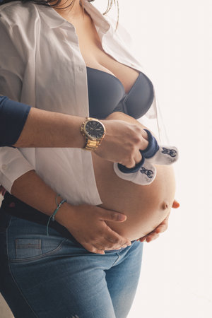 Pregnant young couple with hands on their bellies and holding small shoes on their bellies next to a window with a white backgroundの写真素材