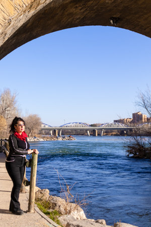 Older latin woman looking at the horizon next to a river in the city of Zaragoza, wearing black clothes, wearing sunglasses and standing under an old bridge.の写真素材