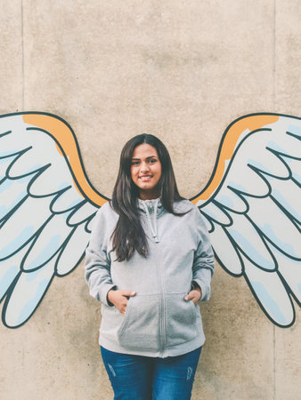 Latin woman looking at the camera and in the background a wall with a drawing of white and yellow wingsの写真素材