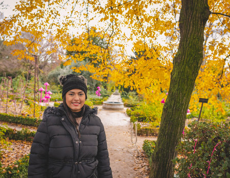 Young Latin woman smiling at the Jardrin Botanico in Madrid, Spain. With yellow and green trees and a water fountain in the background.の写真素材