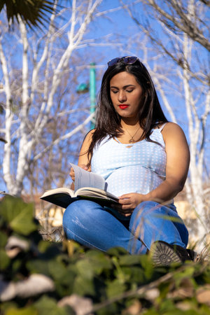 Pregnant Latina woman sitting on the grass, reading a book. She is wearing blue and white clothes with autumn trees in the background.の写真素材