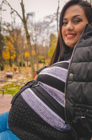 Pregnant woman sitting on a chair in a park, in the city of Madrid, in autumn, wearing dark clothes.の写真素材
