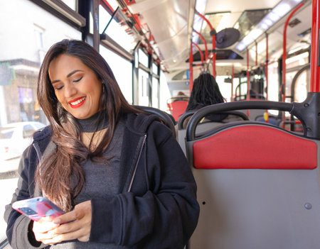 Smiling young woman traveling in an urban bus while looking at her cell phone, red colors predominate on the busの写真素材