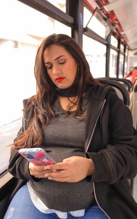 Pregnant Latina woman traveling on an urban bus while looking at her cell phone, red colors predominate on the busの写真素材