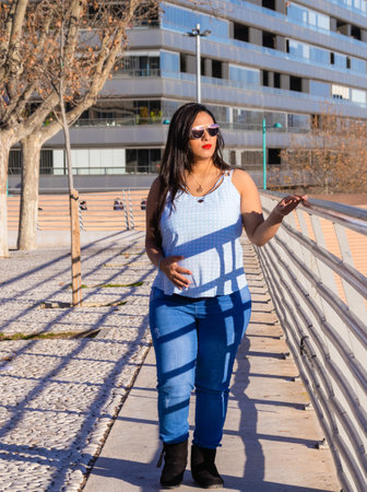Young pregnant woman walking by a fence in the city, looking at the horizon while touching her belly, wearing a blue blouse, jeans and sunglasses.の写真素材