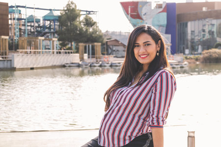 Pregnant Latina woman, sitting on a chair with a lake in the background, sipping and looking at the camera.の写真素材