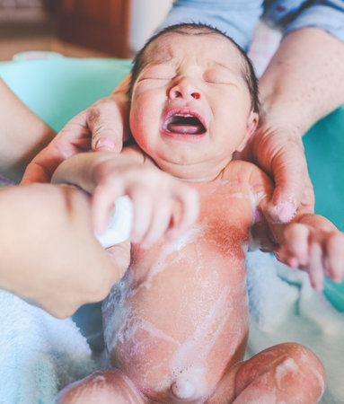 Newborn baby crying in the bathtub while being bathed by two peopleの写真素材