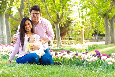 Straight couple with their newborn baby in a park, looking at the camera and tulips in the background.の写真素材