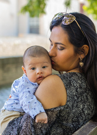 Young latin mom kissing her baby's head, sitting in a park.の写真素材