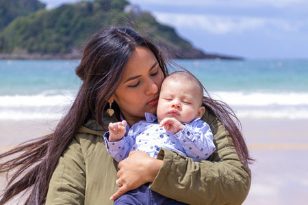 Latin woman tenderly looking at her baby's face, on the beach and in springtimeの写真素材