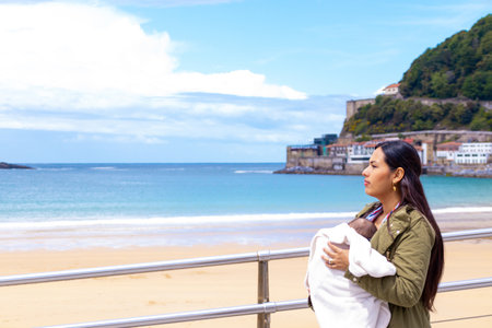 Latin woman on the beach, with her baby in a baby carrier, blue sky and in springtime.の写真素材