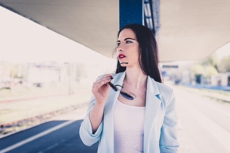 Portrait of a beautiful attractive woman with red lips. Cute girl in a blue jacket and a concentrated pose posing on the platform of a railway station. Female in sunglasses. Retro, vintage style.の写真素材