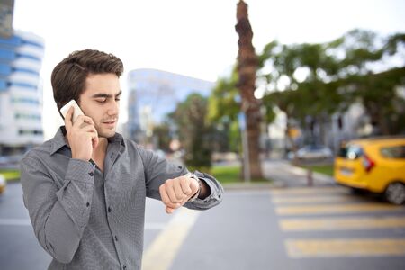 Young businessman talking on mobile phone and  looking at watch on street