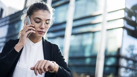 Businesswoman talking mobile phone and looking at watch in the outdoorsの写真素材