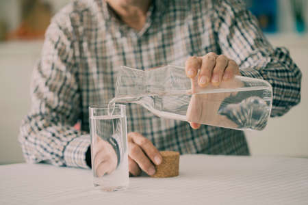 Old man pouring water from bottle to glass in kitchenの写真素材