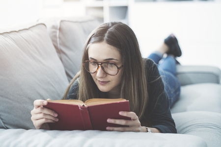 Young girl reading book while lying on couch at homeの写真素材