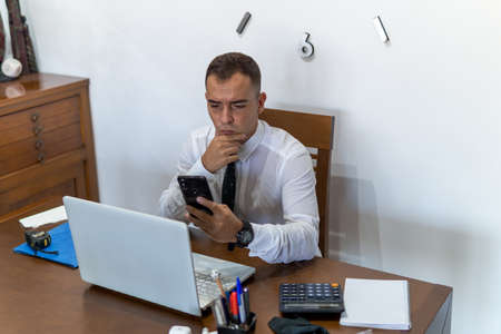 Young businessman working from home office. He is checking his email from his cell phone. In the office he has a laptop computer, a calculator and a notebook.の写真素材