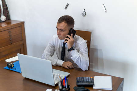 Young businessman working from home office. He is checking his email from his cell phone. In the office he has a laptop computer, a calculator and a notebook.の写真素材