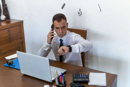 Young businessman working from home office. He is checking his email from his cell phone. In the office he has a laptop computer, a calculator and a notebook.の写真素材