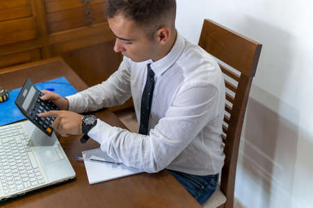 Young businessman working from home office. He is making calculations. In the office he has a laptop computer, a calculator and a notebook.の写真素材