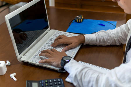 Young businessman working from home office. He is using his laptop. In the office he has a laptop computer, a calculator and a notebook.の写真素材