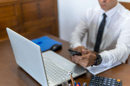 Young businessman working from home office. He is checking his email from his cell phone. In the office he has a laptop computer, a calculator and a notebook.の写真素材