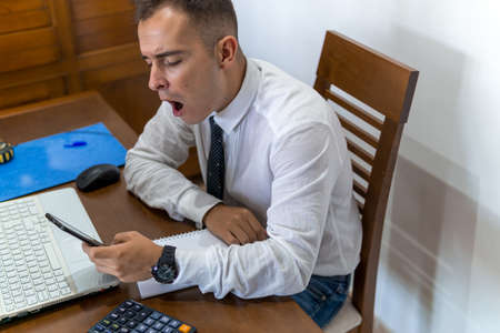 Young businessman working from home office. He is yawning due to fatigue. In the office he has a laptop computer, a calculator and a notebook.の写真素材