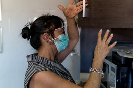Woman searching in kitchen cupboard during covid pandemic. She wears the protective mask.の写真素材
