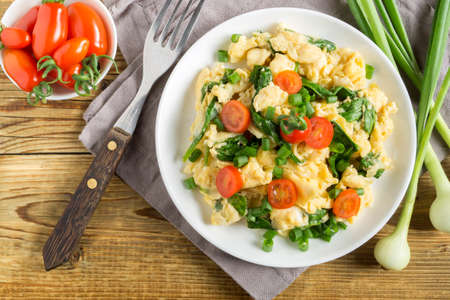 scrambled eggs with spinach and tomatoes on a white plate. on a wooden tableの写真素材