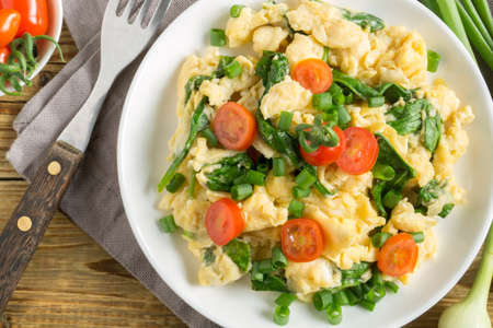 scrambled eggs with spinach and tomatoes on a white plate. on a wooden tableの写真素材