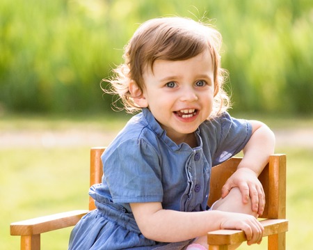 little child, toddler girl in outdoors in the garden sitting on a chair on a sunny summer dayの写真素材