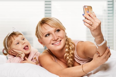 Mother and dauhter taking photo with her smart-phone camera in the bedの写真素材
