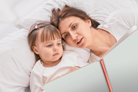 Mother and young daughter reading book on couch at homeの写真素材