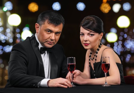 Beautiful young couple with glasses of red wine in luxury restaurant, night lights background