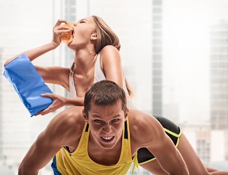 fitness, sport, training, teamwork and lifestyle concept - man starts out push-ups and girl lying on him and eating. on light window backgroundの写真素材