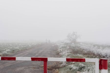 Red and white road barrier on foggy autumn day and poorly distinguishable silhouettes of two peopleの写真素材