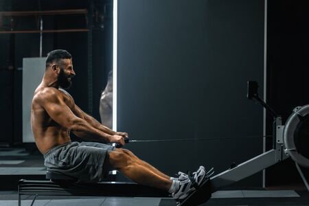 Side view of strong healthy man doing exercises with rowing machine in modern gymの写真素材