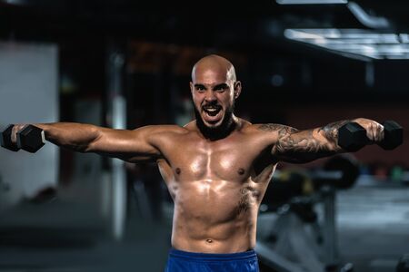 Young fit muscular man doing exercise with dumbbells in gymの写真素材