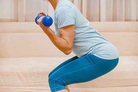 Closeup photo of young athletic girl squatting with dumbbells at home