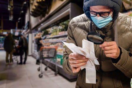 Young man with magnifier inspecting check with grocery purchases during outbreak