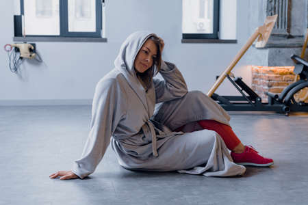 Adult tired sporty woman in gray bathrobe sitting on floor in modern gymの写真素材