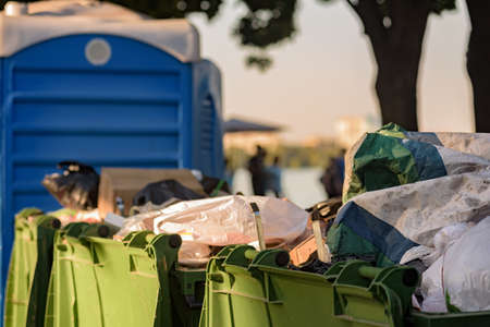 Overflowing waste bins and portable toilet after public event, container for temporarily storing waste, made out of metal or plastic, garbage collection before disposal.の写真素材