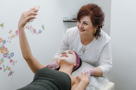 Young woman making selfie with cosmetician after applying facial maskの写真素材
