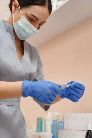 Female cosmetologist wearing protective face mask and gloves and holding syringe for antiaging procedureの写真素材