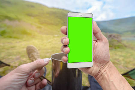 Traveler hiker holding smartphone with green screen in hand in summer mountains
