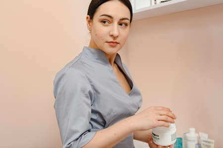 Dnepr, Ukraine- April 20, 2021: Young beautiful cosmetologist in uniform holding skincare cream in beauty salonのeditorial素材