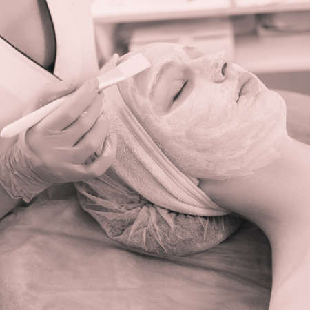 Closeup photo of cosmetologist applying moisturizing mask on female face by brush, monochrome image...